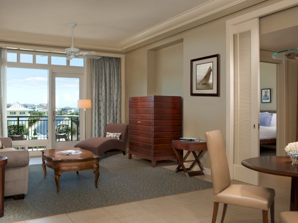 A modern living room with a sofa, chairs, table, and a view to a balcony. Neutral tones and elegant furnishings complete the scene.