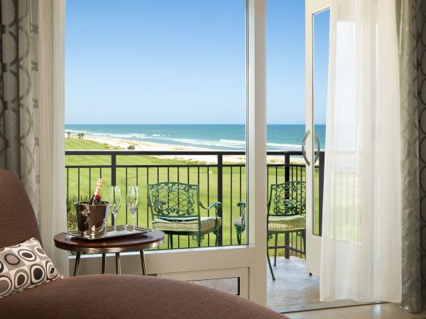 A cozy room overlooks a sunny beach view, with a seating area, curtains, and a table holding a wine set, offering relaxation.