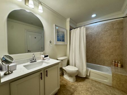 A modern bathroom with a double vanity, large mirror, toilet, and a bathtub/shower combo with beige tile and a shower curtain.