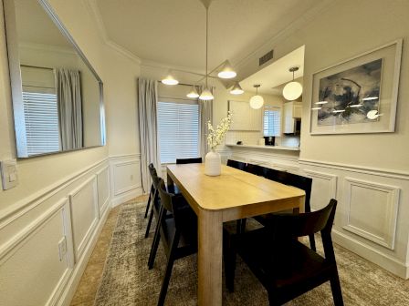 A bright dining room with a wooden table and six chairs, wainscoting on cream walls, a mirror, pendant lights, and a modern art print.