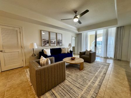 A bright living room with two green armchairs, a navy sofa, a wooden coffee table, a large rug, ceiling fan, and sliding glass doors to a balcony.