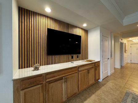 A wood-toned cabinet runs along a wall with a flat-screen TV mounted on a slatted wood backdrop, bright ceiling lights, and a tiled floor ending in a hallway.