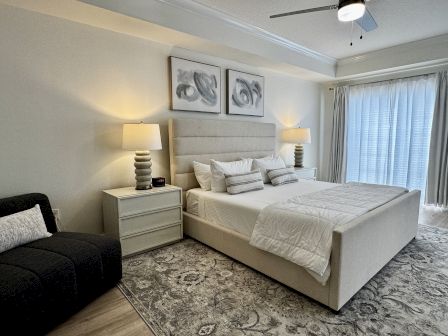A modern hotel-style bedroom with a large beige bed, two matching nightstands, lamps, a dark sofa, and a patterned rug, plus a ceiling fan and sheer curtains.