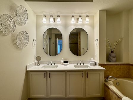 A modern bathroom with dual sinks, two oval mirrors, a light bar above, decorative wall art, and a bathtub to the right.