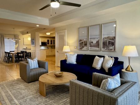 Cozy living room with a navy sofa, striped chairs, neutral pillows, a round wooden coffee table, lamps, and a triptych wall art set, open to the kitchen.