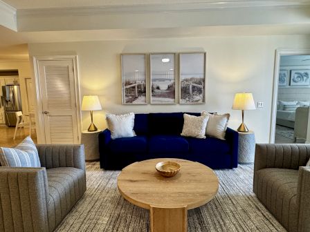 A cozy hotel living room with a navy sofa, two striped armchairs, round wooden table, and warm lamps flanking framed beach artwork.