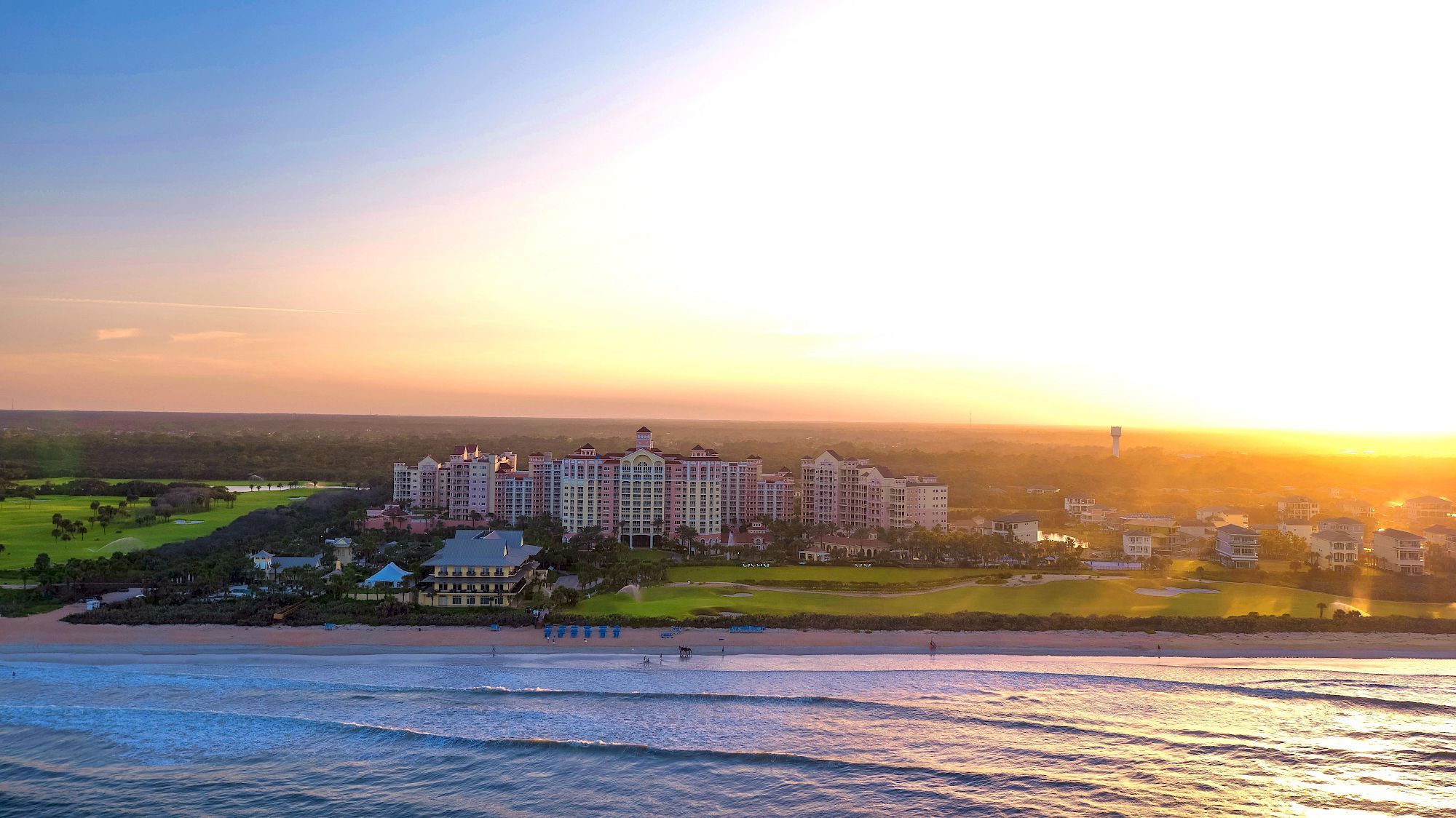 A seaside resort with white buildings sits along a sandy shore while a sunset lights the horizon over calm blue water.