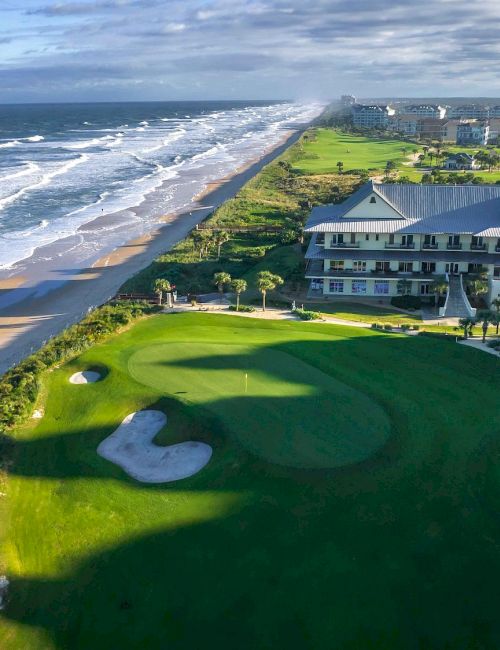 Aerial view of a coastal golf course beside a sandy beach, with a large clubhouse, green fairways, and waves crashing along the shore.