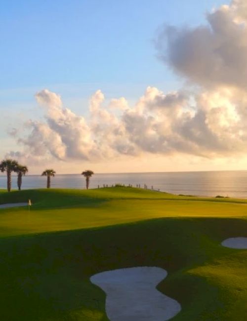 Sunlit coastal golf course with green fairways, sand bunkers, and a calm sea under a dramatic, partly cloudy sky.