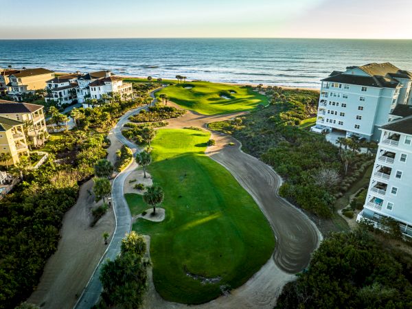 Aerial view of a coastal golf course flanked by hotels, with fairways winding toward the ocean, under a clear sky.