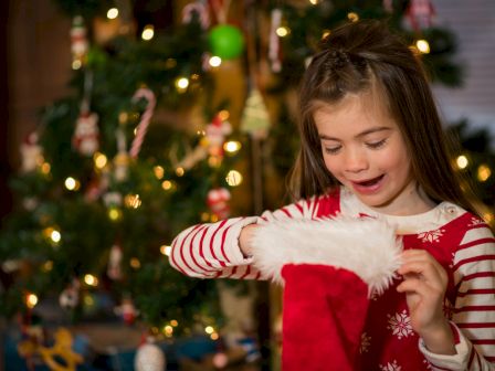 A young girl smiling while inspecting a red Christmas stocking in front of a decorated Christmas tree.