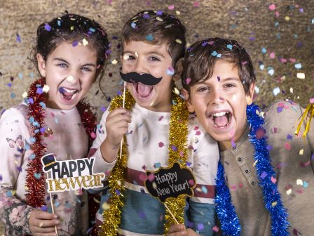 Three kids celebrate, wearing party props and confetti, posing with playful accessories and signs that say &ldquo;Happy New Year&rdquo; in a festive scene.