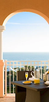 A woman in a white cover-up stands on a terrace overlooking the ocean, while a man in a white shirt sits at a table with breakfast setup.