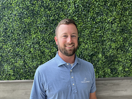 A smiling man in a light blue polo shirt and beige pants stands indoors against a green leafy wall and gray wood paneling.
