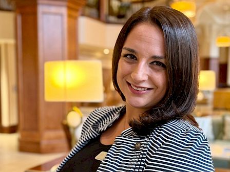 A smiling woman with shoulder-length dark hair wearing a striped blazer in a warmly lit hotel lobby.