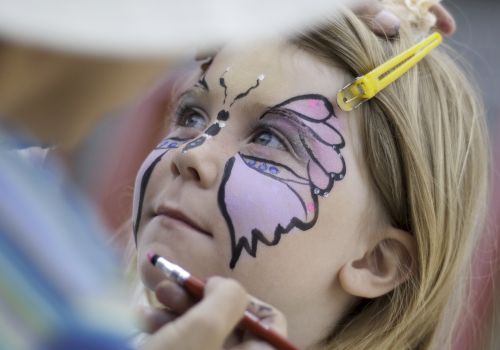 A child gets face-painted like a butterfly by an artist, with colors outlined in purple and pink, focused as makeup is applied.
