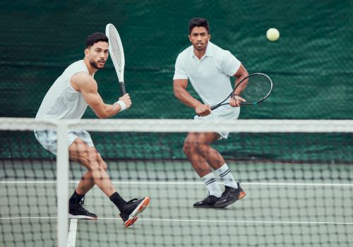 Two men in white shirts play doubles tennis at the net, ready to volley as a ball sails overhead.