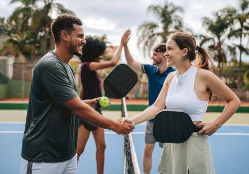 Two people shake hands on a tennis court after a friendly match, smiling while holding paddles, with others celebrating in the background.