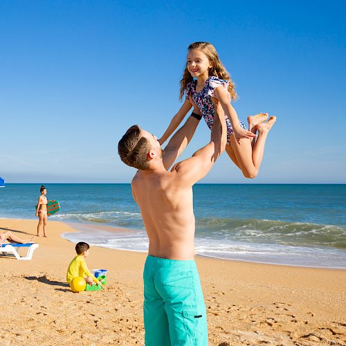 A man lifts a girl in a playful manner on a sunny beach, with others relaxing under blue umbrellas and a child playing with a beach ball.