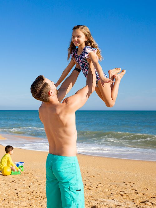 A man lifts a girl in a playful manner on a sunny beach, with others relaxing under blue umbrellas and a child playing with a beach ball.