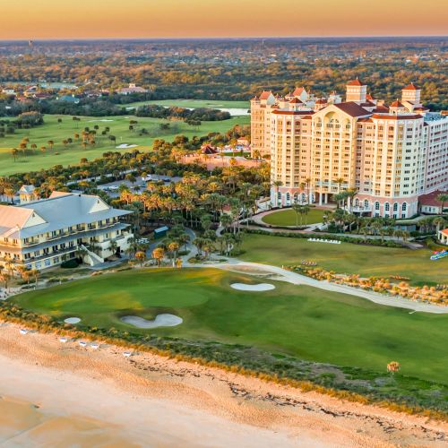 Aerial view of a coastal resort with large buildings, a golf course, and a sandy beach along the ocean.