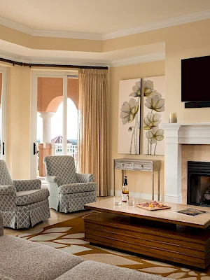A cozy living room with a fireplace, TV, armchairs, sofas, and a balcony view. A coffee table holds wine and glasses.