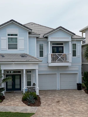 A two-story blue house with a double garage, front porch, small balcony, and a curved brick driveway. The scene looks like a suburban home.