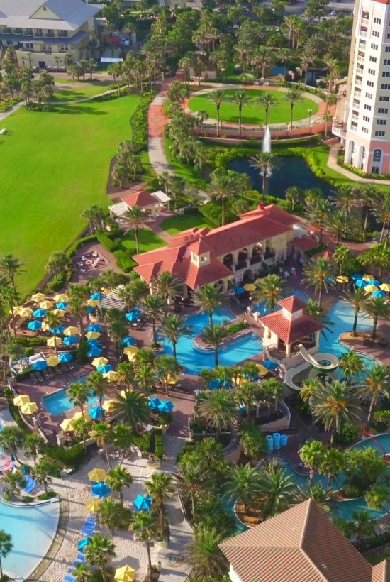 Aerial view of a resort with multiple pools, palm trees, sun loungers, a golf course on the left, and tall beachfront hotel buildings along the shore.