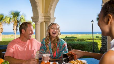 A group of people enjoys a meal and drinks outdoors near the ocean, with palm trees and a clear sky in the background.