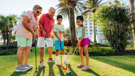 A multi-generational group plays mini-golf outdoors on a sunny lawn with palm trees and buildings in the background.