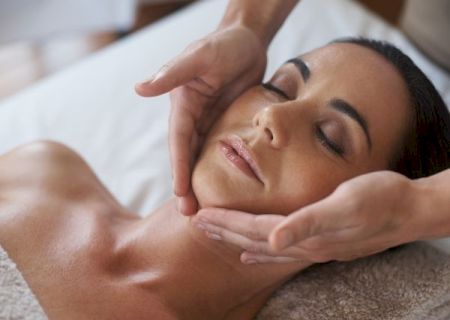 A woman relaxing with a facial massage; hands gently cradle her head and neck as she rests with closed eyes.