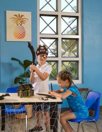 Children are painting at a bright blue room with patterned windows, seated around a table, focused on their craft, a pineapple poster on the wall.