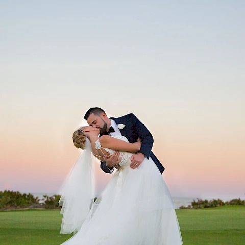 A newlywed couple in wedding attire sharing a kiss outdoors on a grassy field at sunset, with the groom dipping the bride.