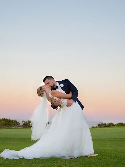 A newlywed couple in wedding attire sharing a kiss outdoors on a grassy field at sunset, with the groom dipping the bride.
