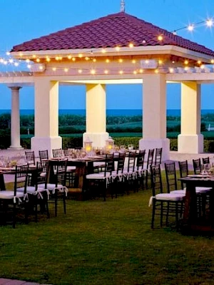 An elegant outdoor wedding reception under string lights around a white pavilion with tables, chairs, and candles at dusk. end.