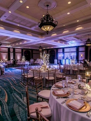 A lavish banquet hall with round tables, gold chairs, elegant place settings, chandeliers, and blue-toned uplighting creating a festive, upscale atmosphere.