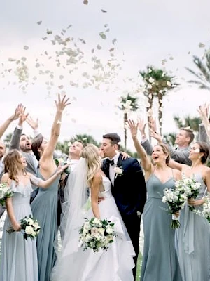 A bride and groom pose with their wedding party as petals scatter above, bridesmaids in blue gowns celebrate with bouquets.