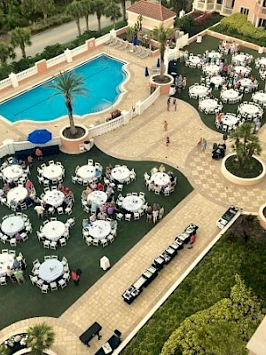 Outdoor wedding setup at a resort: round tables with white linens, chairs, and a long head table, near a pool and palm trees.