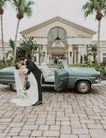 A newlywed couple shares a kiss beside a vintage mint-green car in front of a grand, palm-lined resort entrance, on a stone driveway.