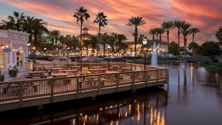 A picturesque waterfront dining area with palm trees, string lights, and a colorful sunset sky reflected in the calm water.