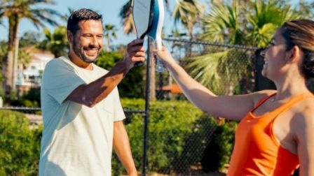 Two people high-fiving a tennis racket on an outdoor court, with palm trees and a clear blue sky behind them.