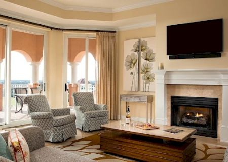A cozy living room with a fireplace, wall-mounted TV, beige walls, large windows, and comfortable seating around a wooden coffee table, balcony doors open.