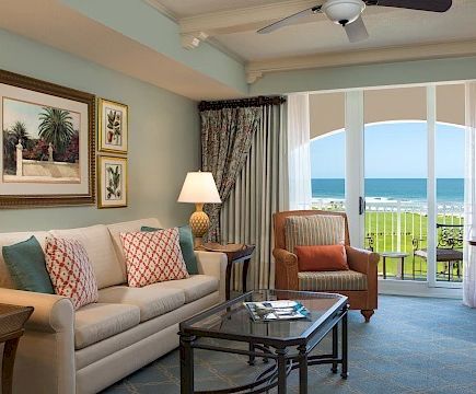 A bright living room with a beige sofa, patterned pillows, two lamps, framed art, and a balcony view of the beach beyond the glass doors.