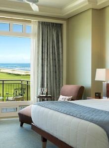 A bright hotel room with a large bed, soft blue bedding, two lamps, a desk chair, and a balcony overlooking a sunny green landscape and ocean beyond.
