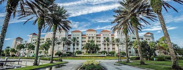A grand resort with white and beige buildings, red-tiled roofs, palm-lined drive, lush green lawns, and a bright blue sky.