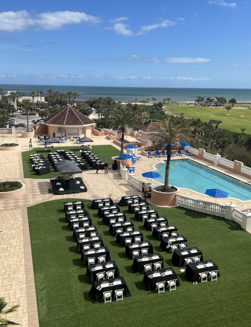 Outdoor event setup with tables and chairs on grass, near a pool and ocean view, under a clear blue sky.