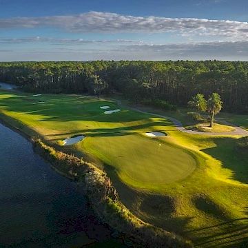 Aerial view of a lush golf course with green fairways, sand bunkers, water hazards, and surrounding trees under a partly cloudy sky.