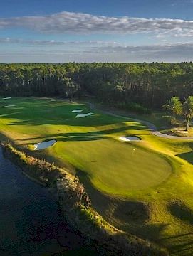 Aerial view of a lush golf course with green fairways, sand bunkers, water hazards, and surrounding trees under a partly cloudy sky.