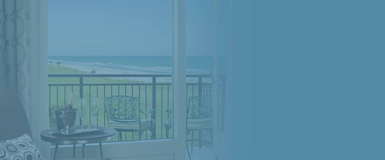 A beach view from a room with a small table, champagne, and chairs on a balcony is shown.