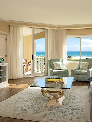 The image shows a luxurious living room with elegant furniture, a coffee table, TV, and a stunning ocean view through large windows.
